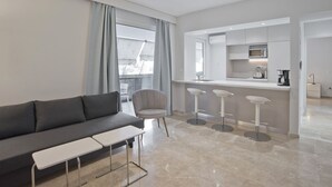A modern living room with a grey sofa, a small white table, and a grey armchair. There is a kitchen area with a microwave, a coffee maker, and a sink. The room has a large window with grey curtains.