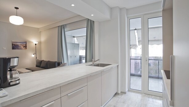 A modern kitchen with a large island, a built-in oven, and a sink. There is a window with curtains and a view of the outside.