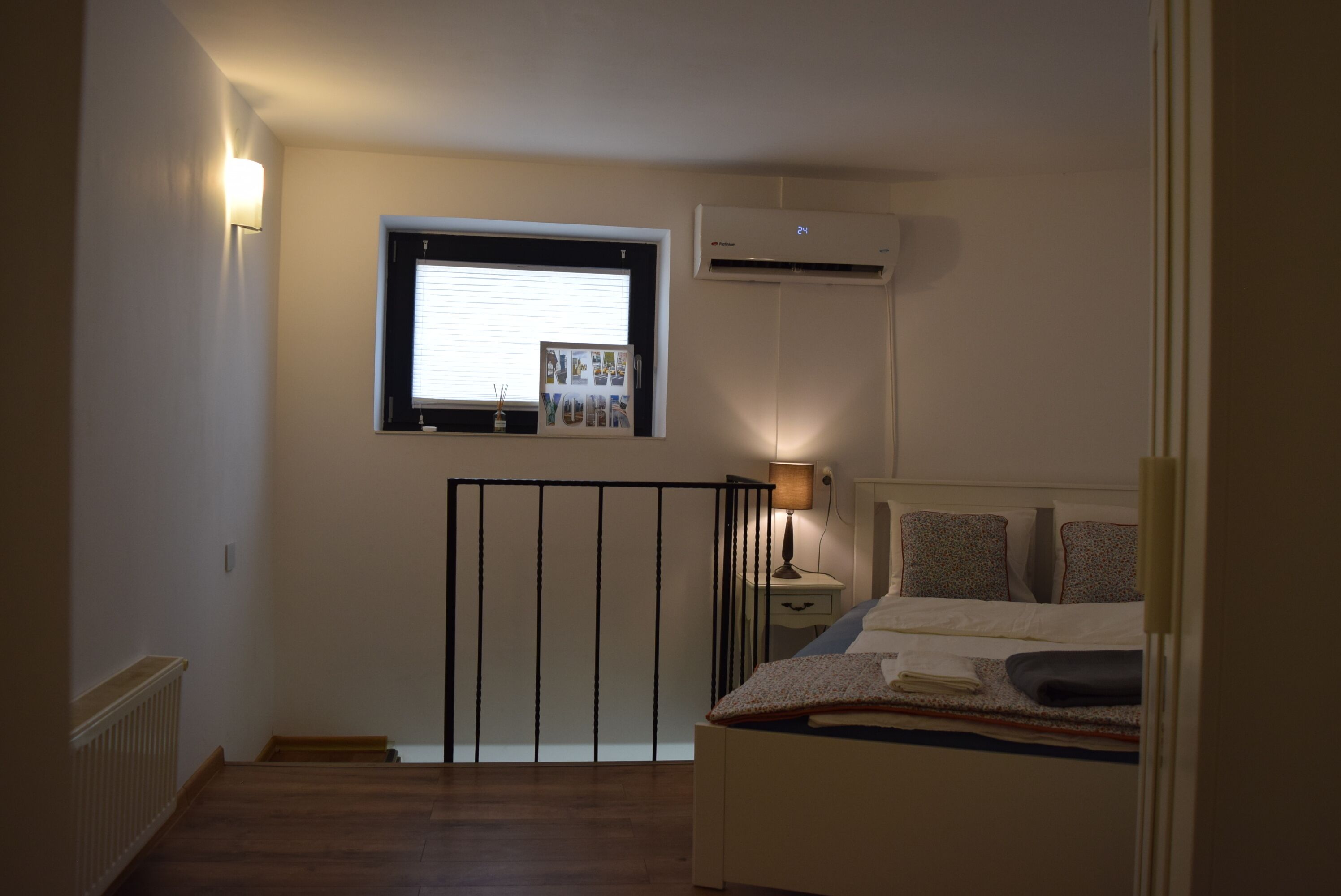 Quarto com cama, janela com persianas, ar condicionado e mesa de cabeceira com luminária.