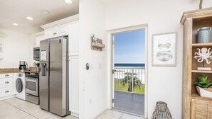 Une cuisine moderne équipée d’appareils électroménagers en acier inoxydable, d’un lave-linge et offrant une vue sur l’océan grâce à une porte-fenêtre donnant sur un balcon.
