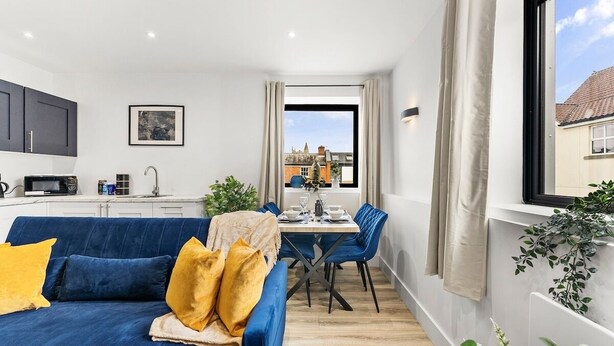 Ein modernes Apartment mit einem blauen Sofa, einem Esstisch und Stühlen. Es gibt eine Küchenzeile mit Schränken und einer Mikrowelle. Ein Fenster bietet einen Blick ins Freie.