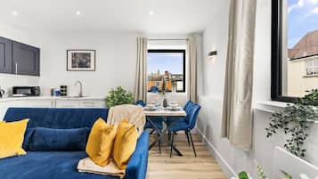 A modern apartment with a blue sofa, dining table, and chairs. There is a kitchen area with cabinets and a microwave. A window provides a view of the outside.