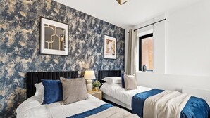 Ein Hotelzimmer mit zwei Betten, einer strukturierten Wand, gerahmten Kunstwerken, einem Fenster mit Vorhängen und einem Nachttisch mit einer Lampe.