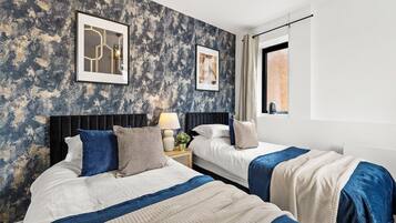 A hotel room with two beds, a textured wall, framed artwork, a window with curtains, and a nightstand with a lamp.