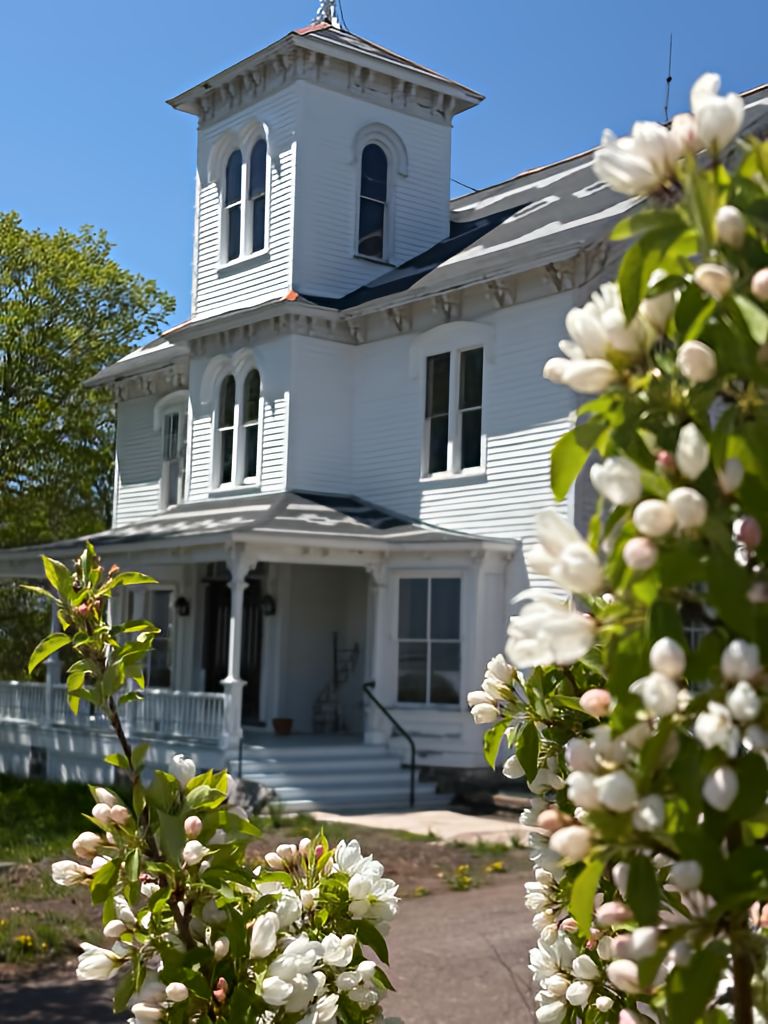 A two-story white building with a central tower, a porch, and a garden with blooming flowers.