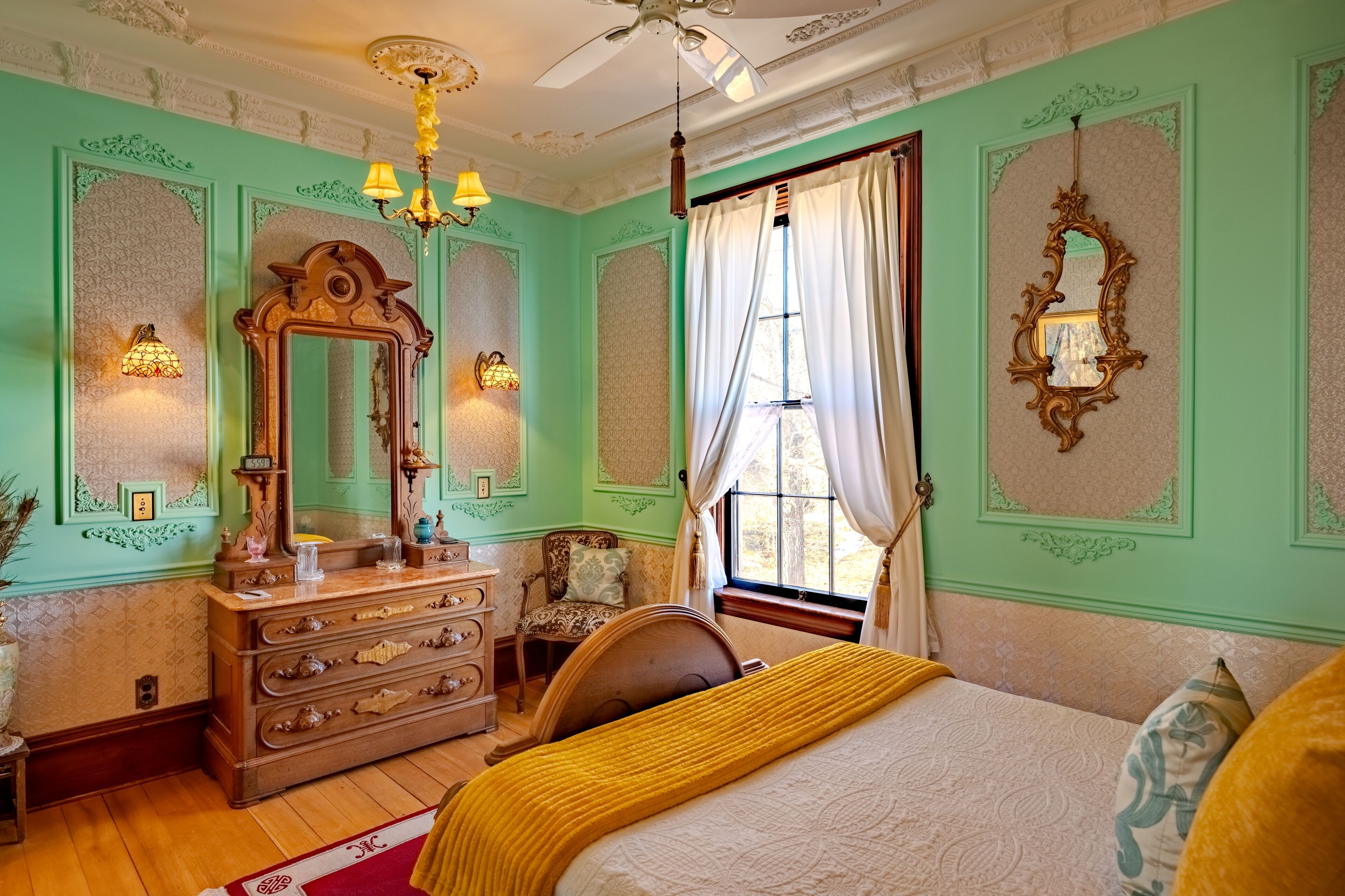 Een kamer met een bed, een kaptafel, een spiegel, een kroonluchter en een raam met gordijnen.