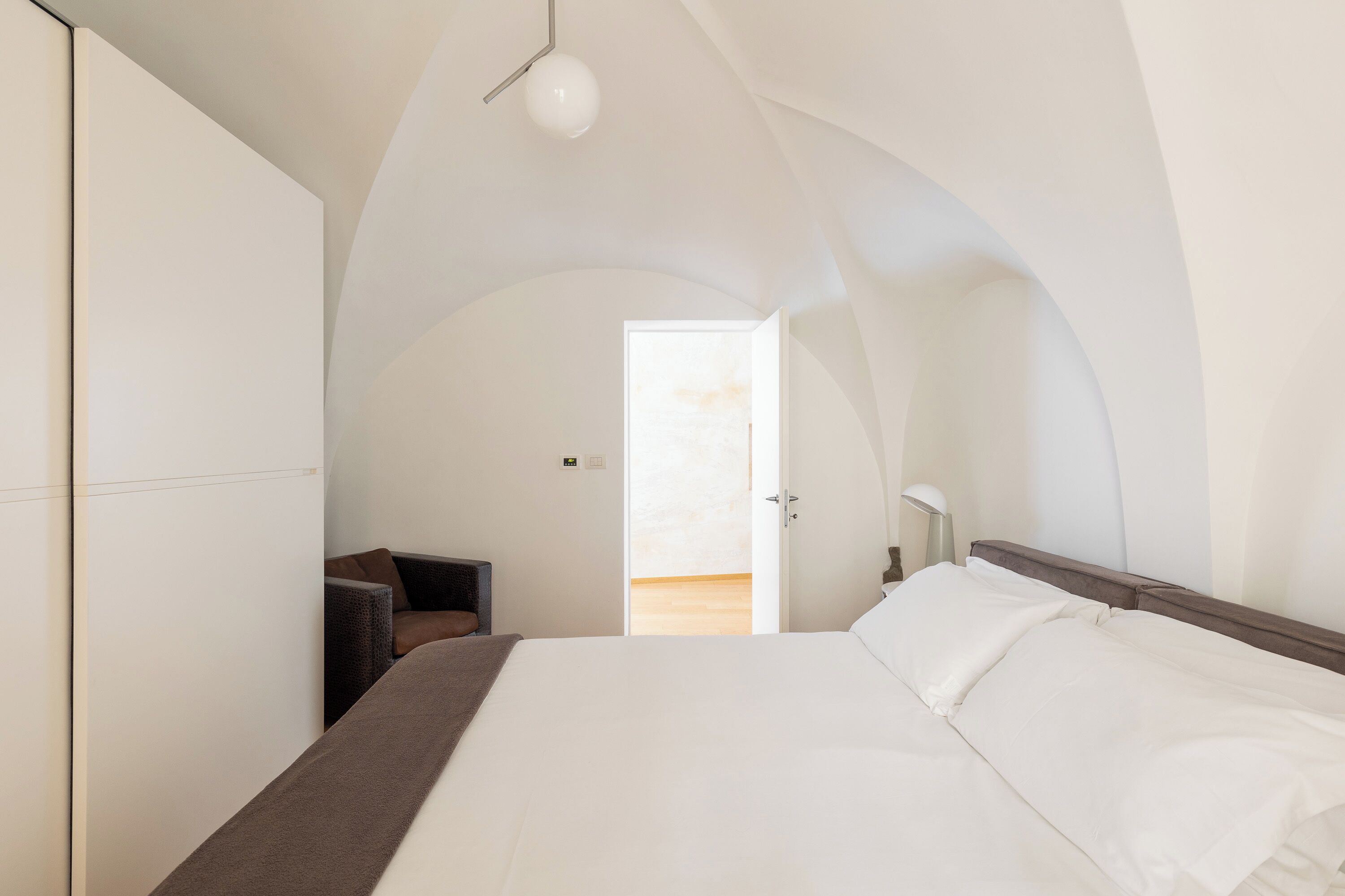 A bedroom with a large bed, a chair, and a desk. There is a door leading to a balcony.