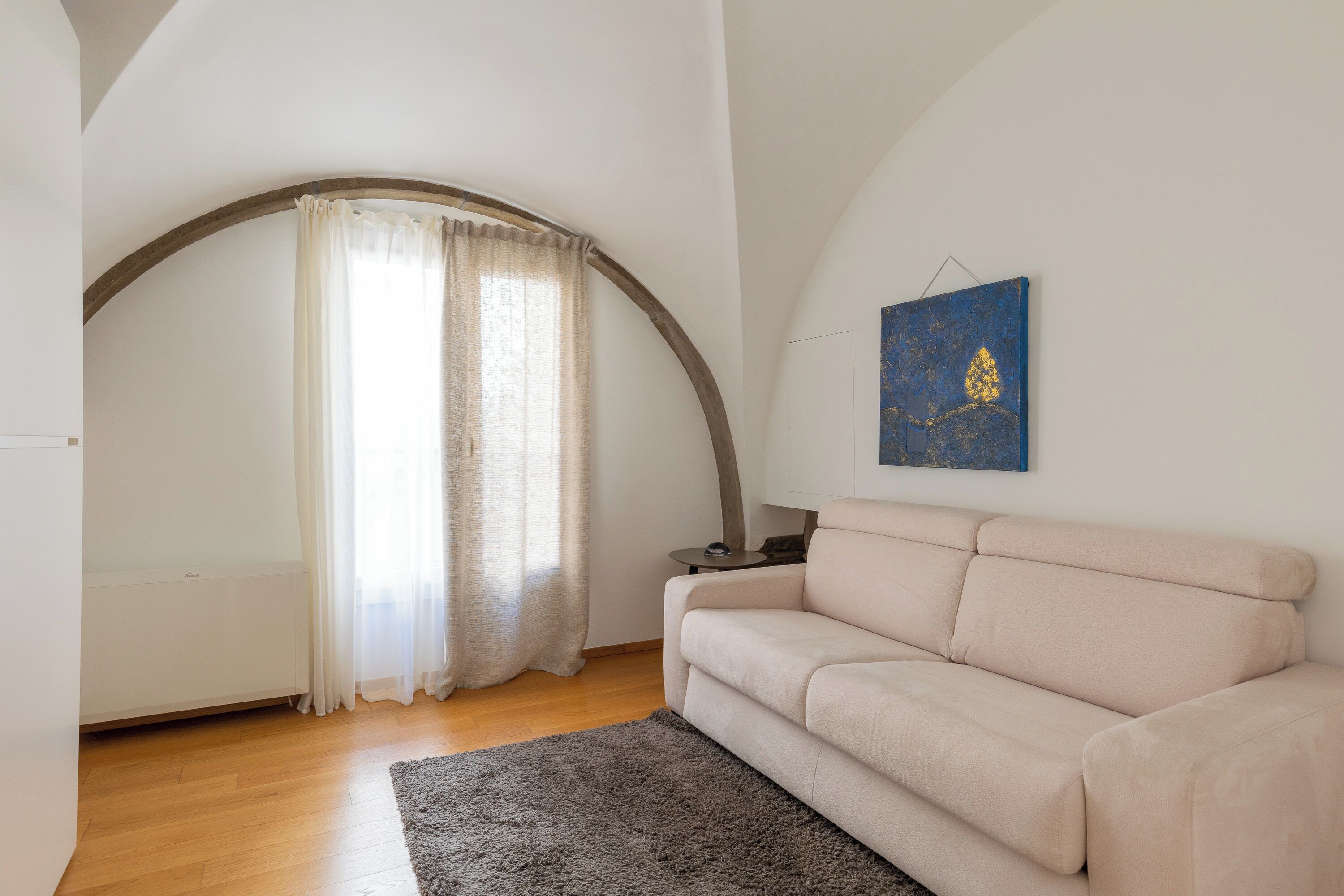 Ein Wohnzimmer mit einem beigefarbenen Sofa, einem grauen Teppich, Holzdielen, einem weißen Heizkörper und einem blauen Kunstwerk an der Wand.