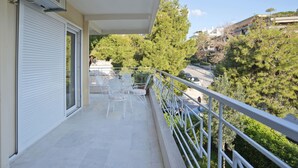 A balcony with a white railing, a table with chairs, and a view of trees and buildings. - Riviera Living in Vouliagmeni (Vari-Voula-Vouliagmeni)