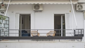 Ein Balkon mit zwei Korbstühlen und einem kleinen Tisch, einer weißen Außenwand und zwei offenen Türen, die ins Innere führen.
