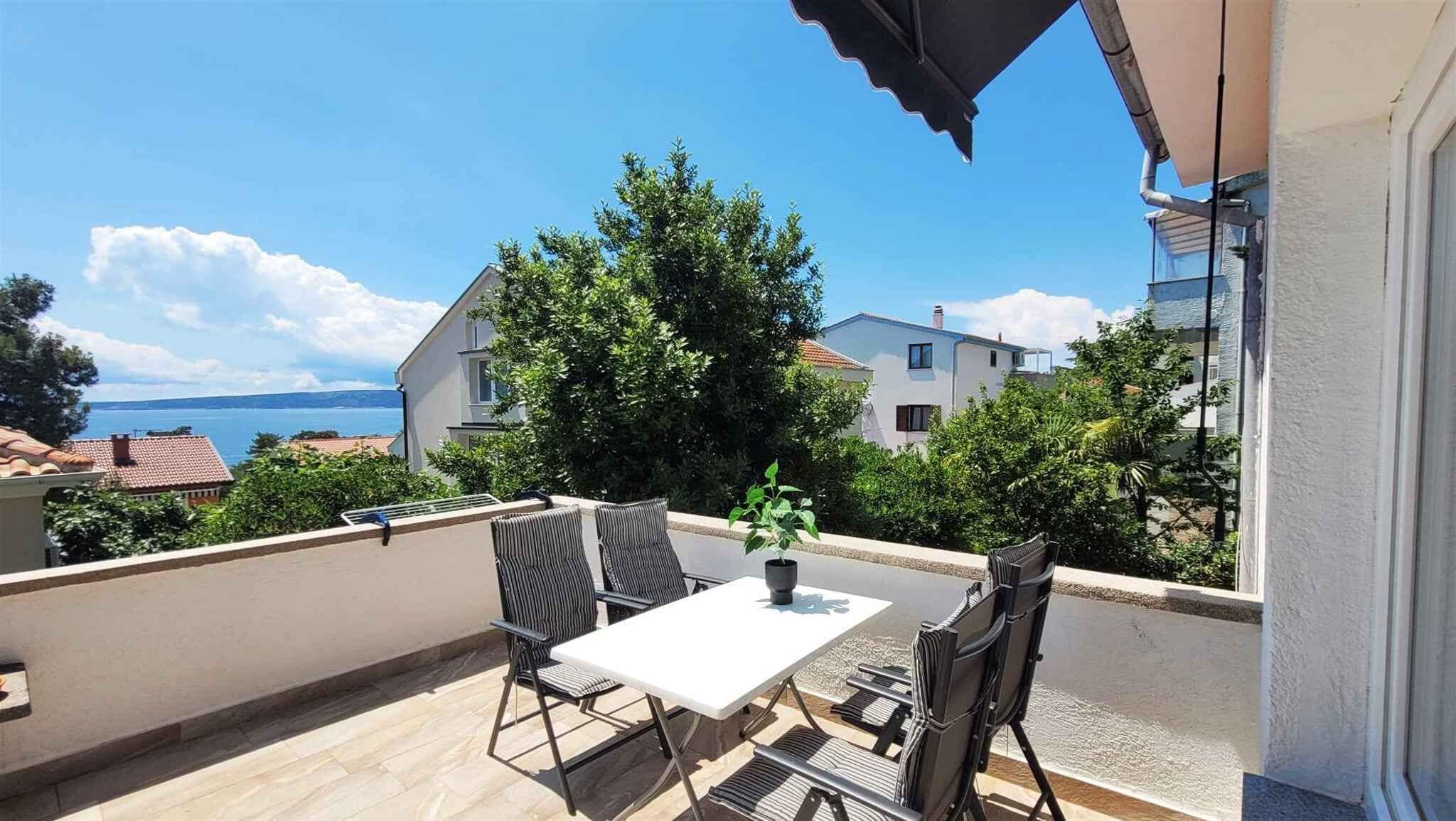 A balcony with a table and chairs, a potted plant, and a view of the sea and buildings.