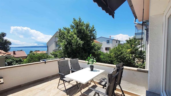 A balcony with a table and chairs, a potted plant, and a view of the sea and buildings.