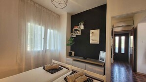 Ein Schlafzimmer mit einem Bett, einem Nachttisch, einer Wandregal mit Büchern und einer Pflanze sowie einem Fenster mit durchscheinenden Vorhängen.