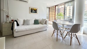 Un soggiorno moderno con un divano bianco, un tavolo da pranzo con sedie e una grande finestra vista edifici.