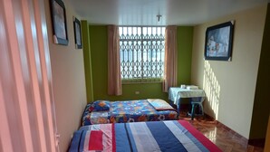 Een kamer met twee bedden, een raam met metalen tralies, een klein tafeltje met tafelkleed en een foto aan de muur.