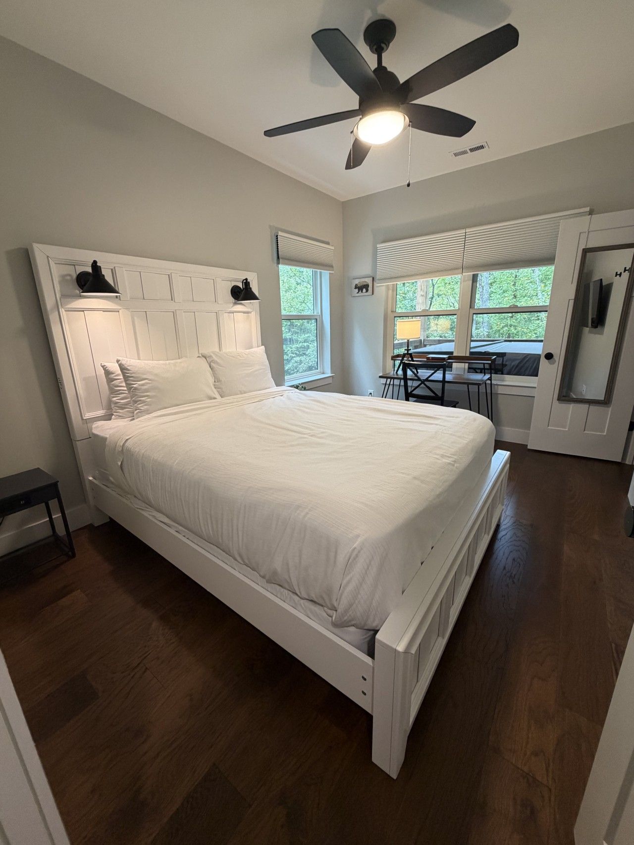 A bedroom with a white bed, a ceiling fan, and a view of the outdoors through windows.