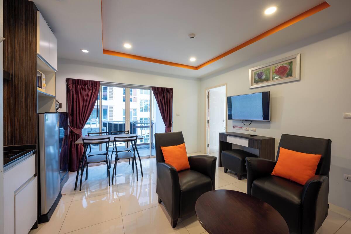 Una sala de estar moderna con comedor, televisor montado en la pared y zona de cocina con refrigerador.