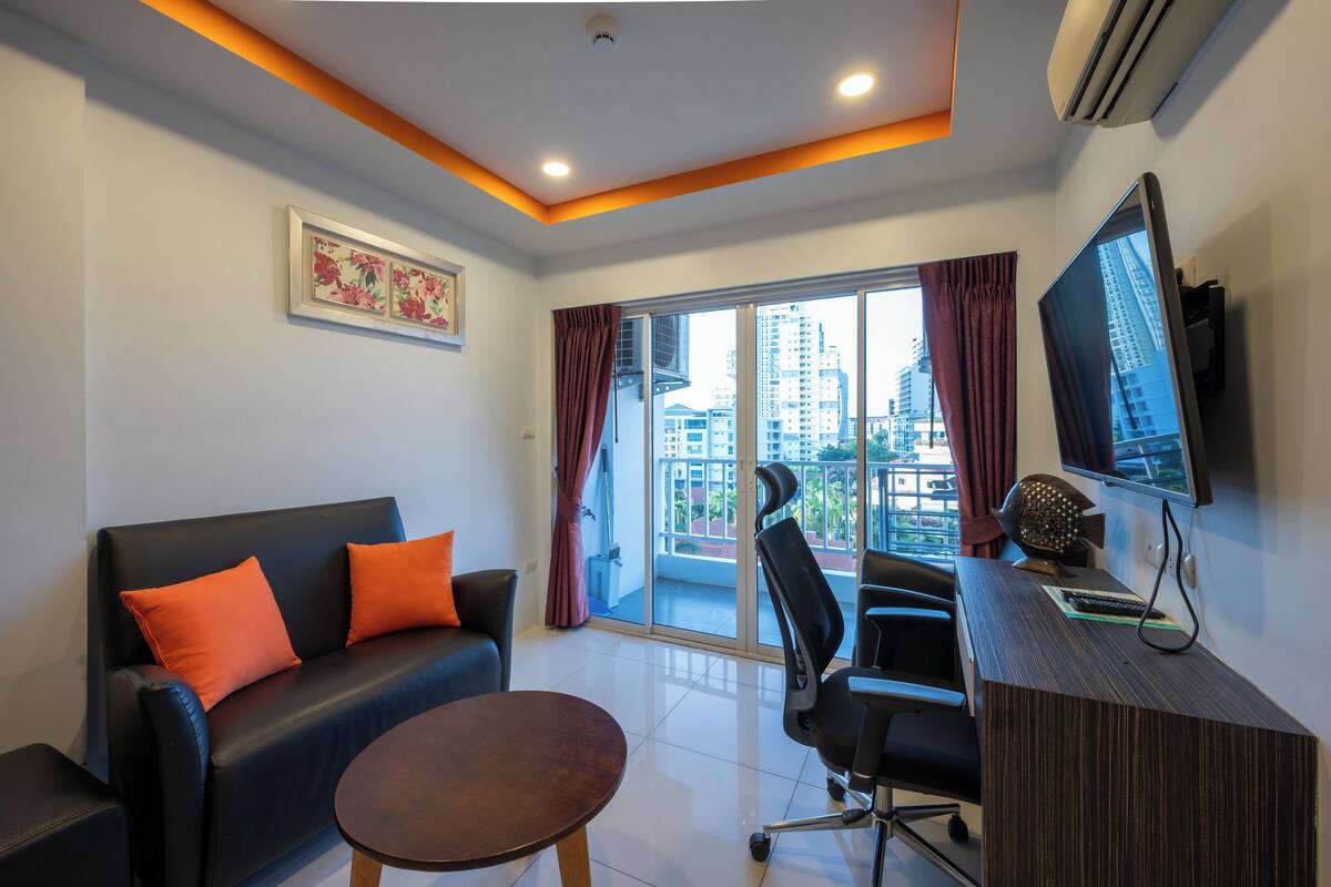 Una sala de estar moderna con un sofá negro, cojines naranjas, una mesa de centro de madera, un escritorio con computadora y un ventanal con cortinas.