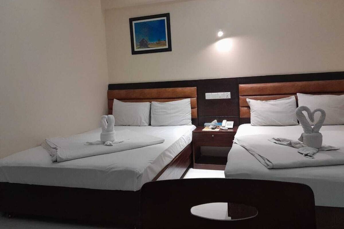 A hotel room with two beds, each with a towel and a heart-shaped towel decoration. There is a nightstand with a phone and a small table between the beds. A picture hangs on the wall above the beds.