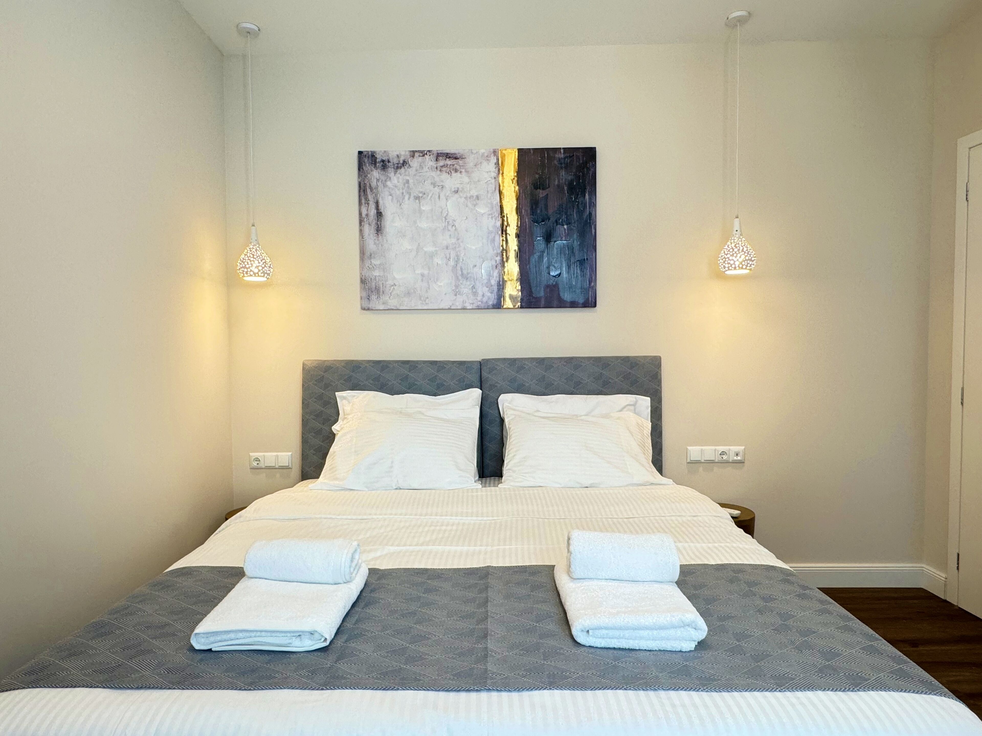 A double bed with a grey headboard and white bedding, two bedside tables, and a modern abstract painting on the wall.