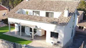 Ein zweistöckiges Haus mit Ziegeldach, einer überdachten Terrasse mit Gartenmöbeln und einer Steinmauer, die das Grundstück umgibt.