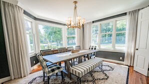 Ein Esszimmer mit großem Tisch, Stühlen und Bänken, umgeben von großen Fenstern und einem gemusterten Teppich.