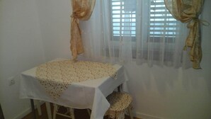 Ein kleiner Raum mit einem Tisch, der mit einer blumigen Tischdecke bedeckt ist, einem Hocker und einem Fenster mit transparenten Vorhängen.