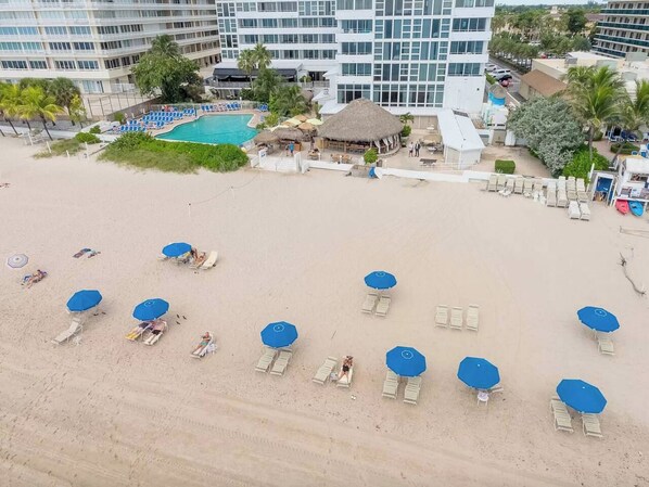 An aerial view of a beach with sun loungers and umbrellas, a pool, and a resort building.