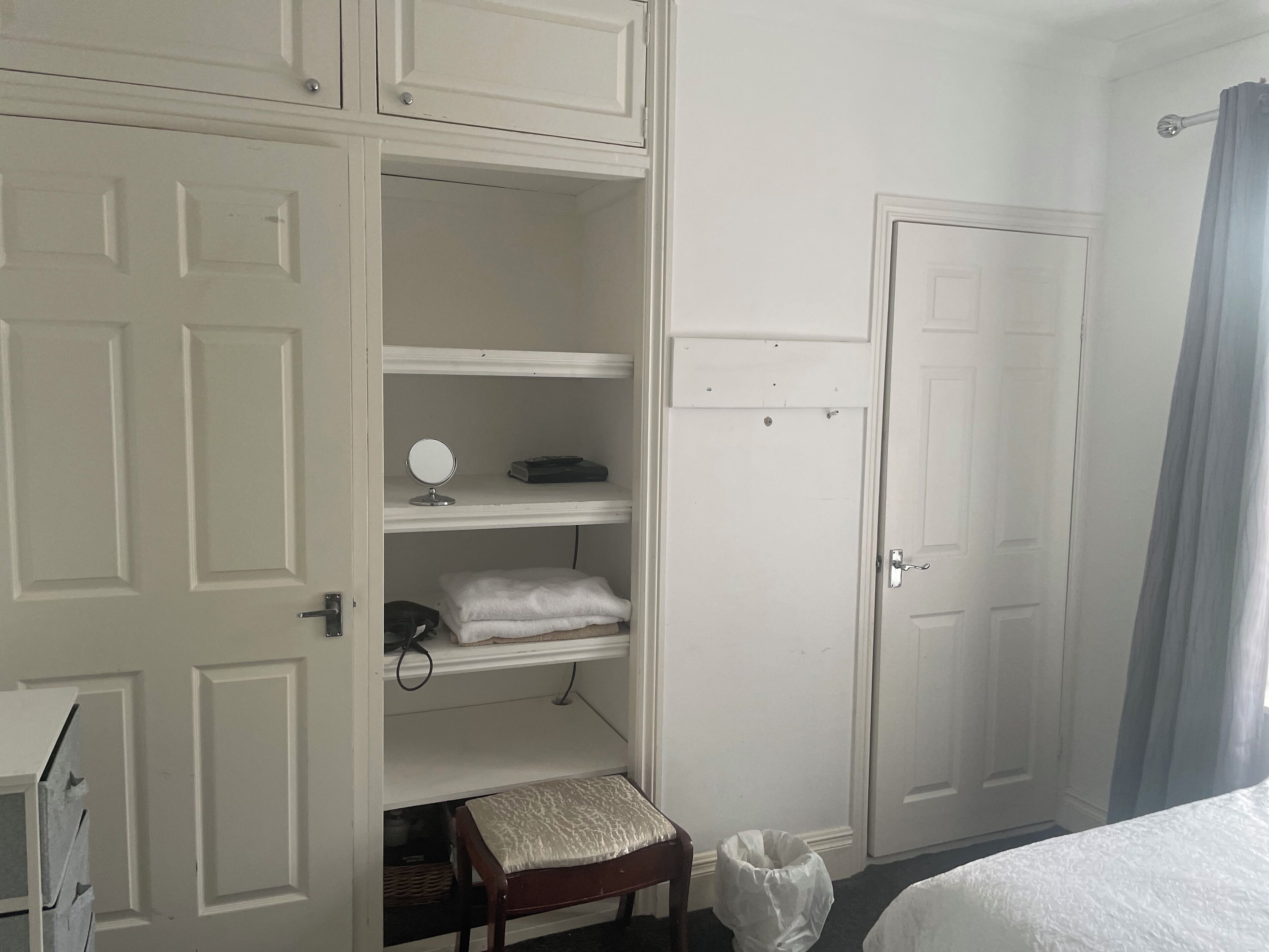 A bedroom with a bed, a small wooden bench, a white trash bin, and a door.