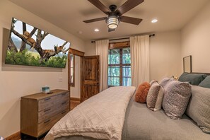 Une chambre à coucher comprenant un lit, une commode, un ventilateur de plafond, une fenêtre avec des rideaux et un téléviseur fixé au mur.