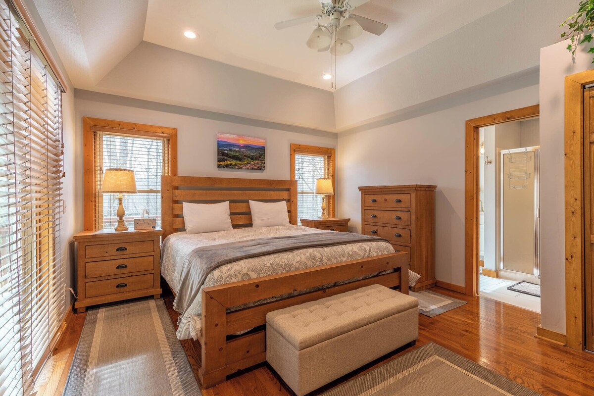 A bedroom with a wooden bed, bedside tables, a bench, and a view of the outdoors through windows.