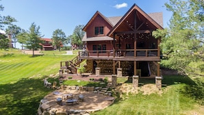 A large, two-story wooden cabin with a stone foundation, a balcony, and a stone fire pit area. - The Sheltered Elk at Eagles Nest (Banner Elk)