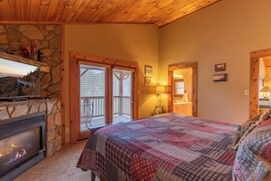 A cozy bedroom with a stone fireplace, a large bed with a plaid quilt, and a scenic view through the door. - Ambleside (Vilas)