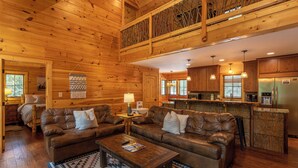 A spacious wooden cabin interior with a living area featuring two leather sofas, a coffee table, and a patterned rug. A kitchen with wooden cabinets and stainless steel appliances is visible in the background. - Laurel Creek Lodge (Banner Elk)