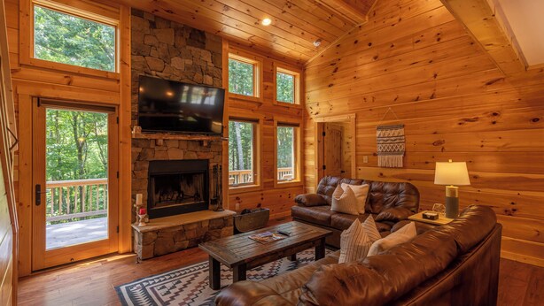 A log cabin interior with a stone fireplace, leather sofas, a flat-screen TV, and large windows. - Laurel Creek Lodge (Banner Elk)