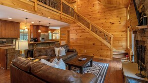 A spacious log cabin interior with a living area featuring a leather sofa, a coffee table, and a fireplace. A wooden staircase with a railing leads to an upper level. - Laurel Creek Lodge (Banner Elk)