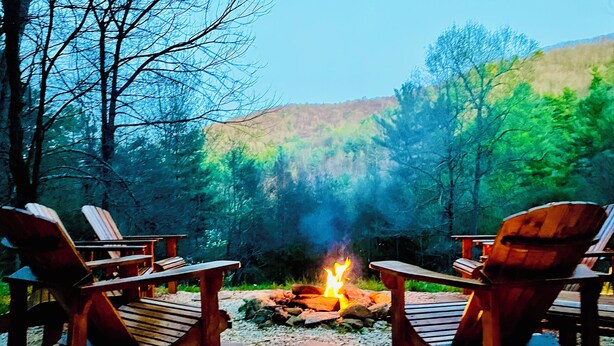 A campfire with two Adirondack chairs facing it, surrounded by a forest. - Moonlight Lodge (Fleetwood)