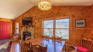 L’intérieur d’un chalet en bois comprend une table à manger, des chaises, une cheminée, un téléviseur et une grande fenêtre donnant sur la forêt.