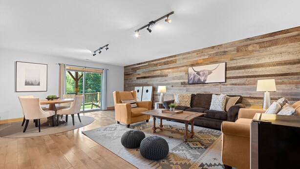 Un salon moderne avec un mur d’accent en bois, un coin repas doté d’une table ronde et de chaises, et une grande fenêtre offrant une vue sur la verdure.
