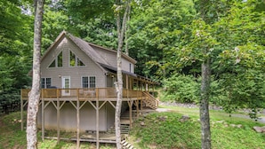 A two-story wooden cabin with a deck, surrounded by trees and a fire pit area. - Gone Ski'Inn (Banner Elk)