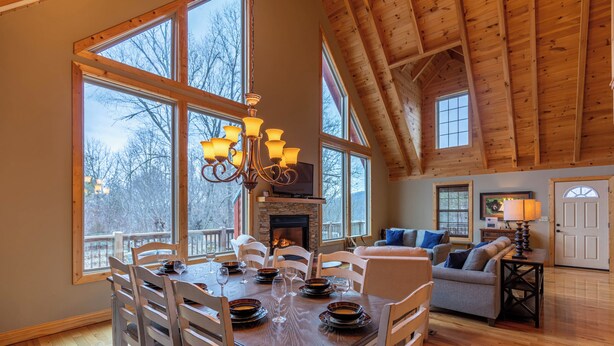 Un coin repas avec une table en bois dressée pour six personnes, entourée de chaises blanches, d’un lustre et d’une cheminée.