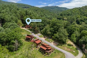 Un chalet doté de plusieurs cabanes en bois, entouré d’une forêt dense, avec une route sinueuse et une chaîne de montagnes en arrière-plan.