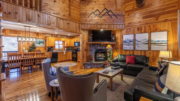 A cozy cabin interior with a wooden interior, a fireplace, a dining area, and a living room with a sofa and a television. - Riverbend Retreat (Todd)