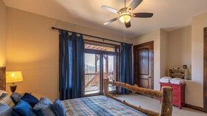 Une chambre à coucher avec un lit, une tête de lit en bois, un ventilateur de plafond, une fenêtre avec des rideaux, une porte en bois et une petite commode rouge.