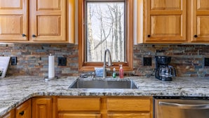 A kitchen with wooden cabinets, a stone backsplash, a window with a view of trees, a stainless steel sink, and a coffee maker. - A Change of Scenery at Yonahlossee (Boone)