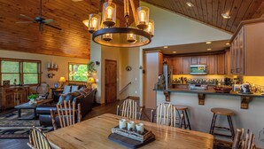 A spacious living area with a wooden ceiling, a dining table with chairs, a sofa, and a kitchen with wooden cabinets and a bar area. - Bearadise Cabin (Banner Elk)