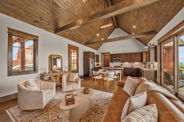 A spacious living area with a high ceiling, wooden beams, a large sofa, a coffee table, and a view through large windows. - Cloud Catcher at Eagles Nest (Banner Elk)