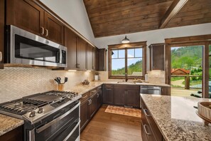 A kitchen with dark wooden cabinets, stainless steel appliances, a herringbone tile backsplash, and a view of a green landscape through a large window. - Cloud Catcher at Eagles Nest (Banner Elk)