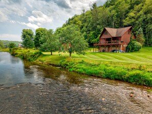 A log cabin with a porch, surrounded by lush greenery and a river. - River Roost (Todd)