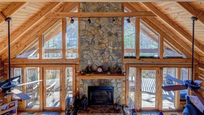 A log cabin interior with a stone fireplace, large windows, and wooden beams. - Fox's Lair (Sugar Grove)